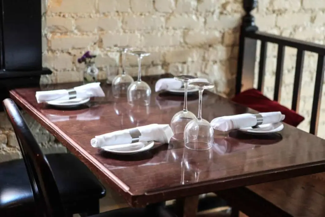 A wooden table set for four with white napkins, plates, and upside-down wine glasses in a restaurant with a textured brick wall.