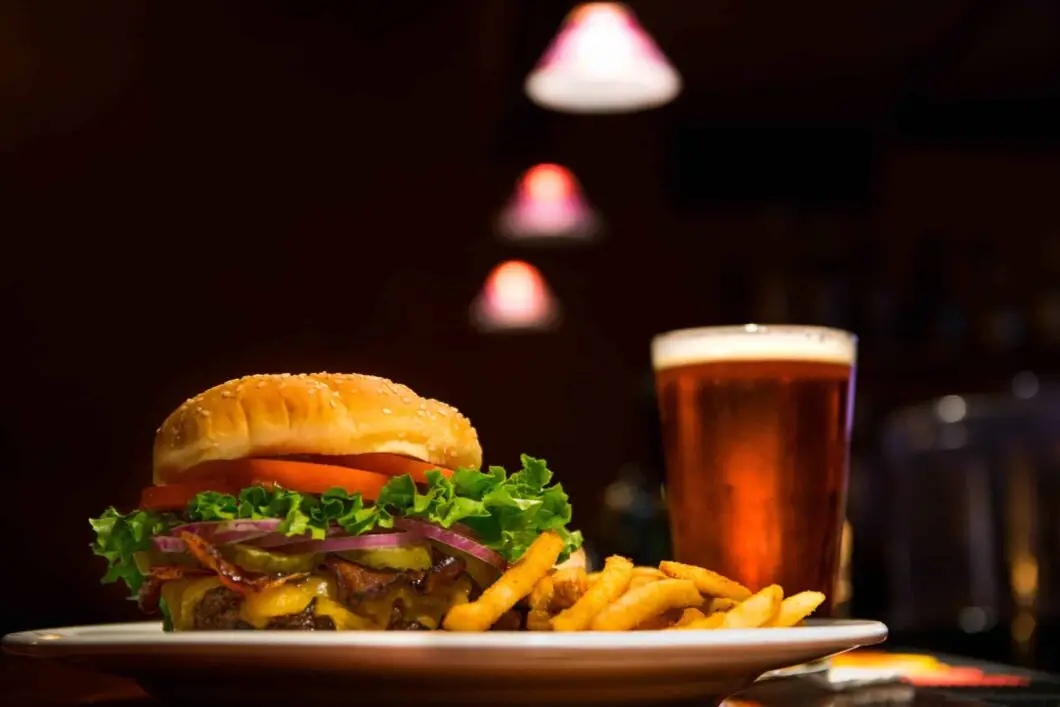A cheeseburger with lettuce, tomato, onions, and fries on a plate next to a glass of beer, with blurred lights in the background.