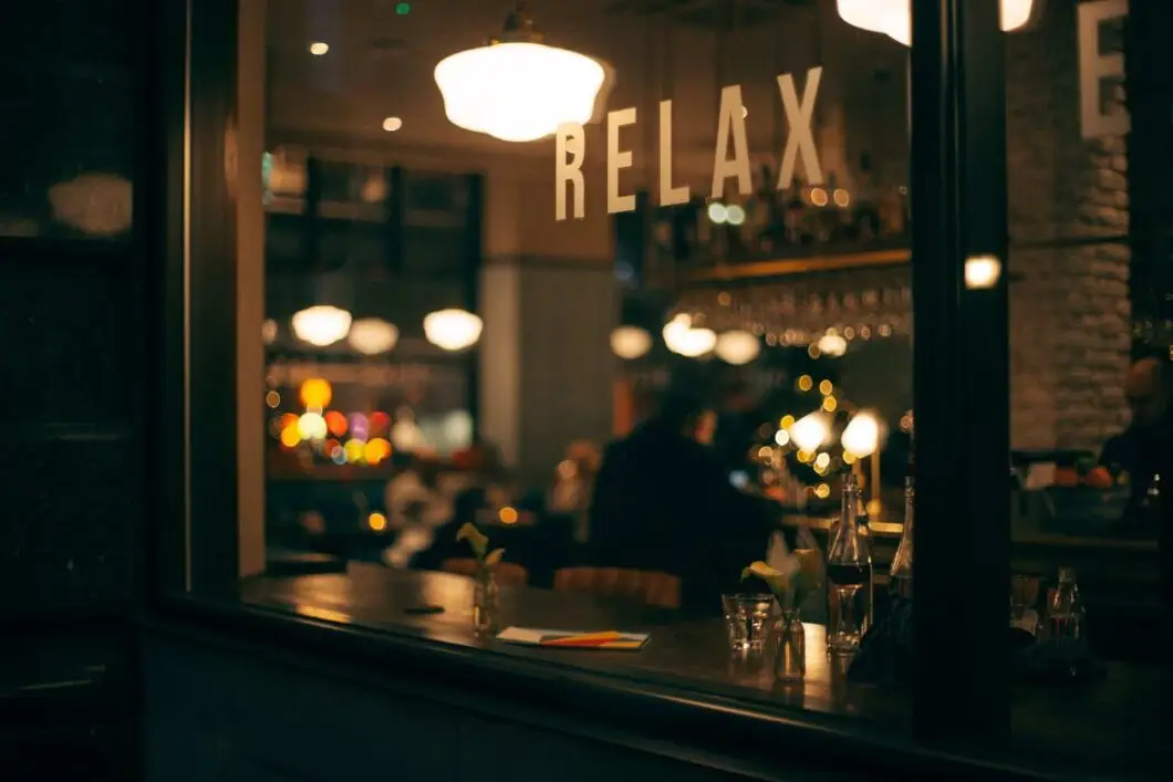 View through a window into a dimly lit bar or restaurant with “RELAX” on the glass; empty glasses and bottles are on a counter, and people are blurred in the background.