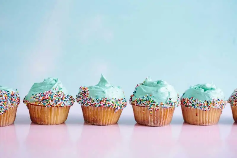 Six vanilla cupcakes with blue frosting and rainbow sprinkles are arranged in a row on a glossy pink surface against a light blue background.