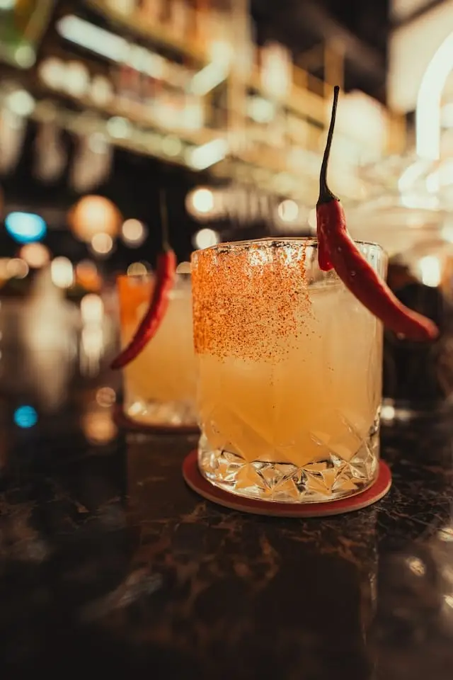 Two cocktails with chili peppers as garnish and chili powder on the rim, served on a dark bar counter with blurred lights in the background.