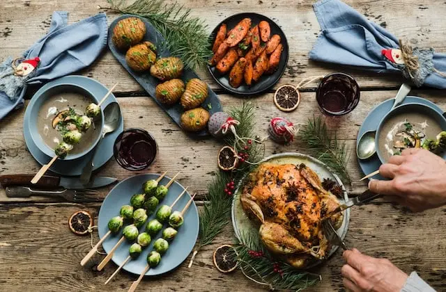 A person carves a roasted chicken on a rustic wooden table set with potatoes, carrots, Brussels sprouts, soup, drinks, and festive decorations.
