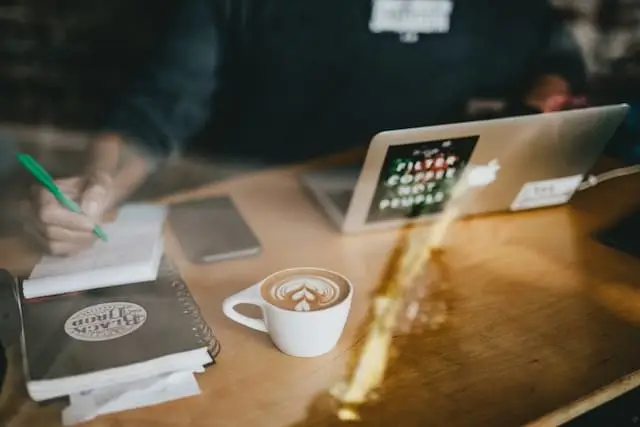 A person writes in a notebook next to a laptop, a smartphone, and a cup of coffee with latte art on a wooden table.