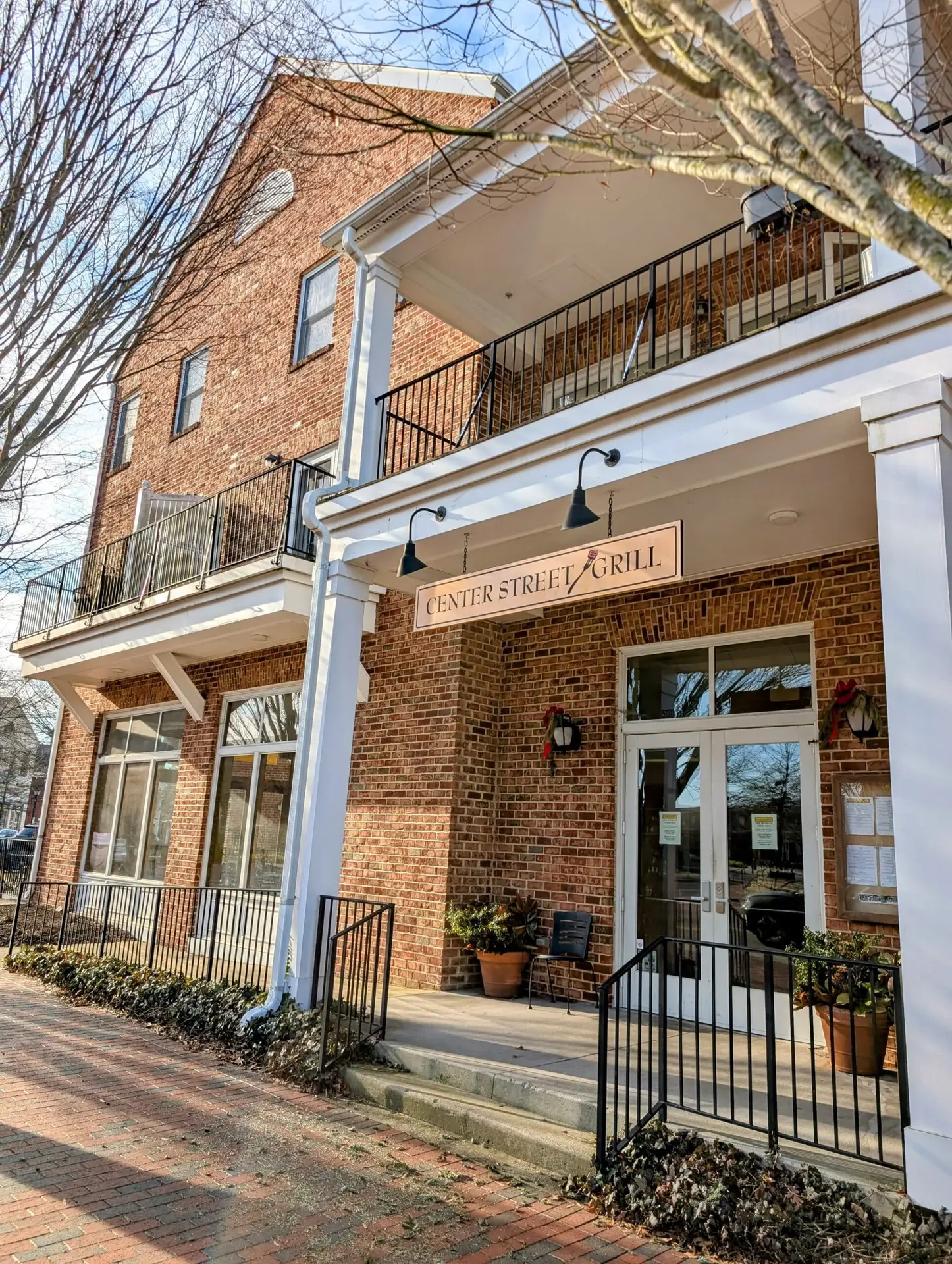 A brick building with a sign reading “Center Street Grill” above the entrance, with potted plants by the door and black railings along the steps and ramp.