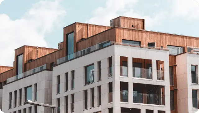Modern apartment building with white facade, large windows, and upper sections featuring wood paneling under a partly cloudy sky.