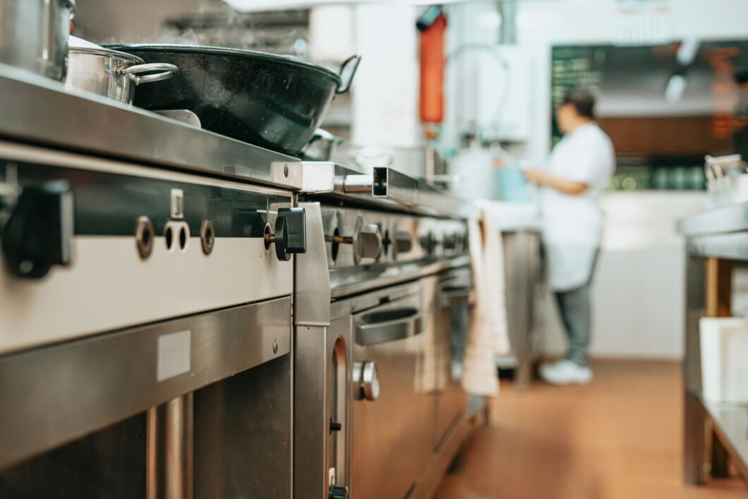 Commercial kitchen with stainless steel range and cookware in foreground, cook working at prep station in background