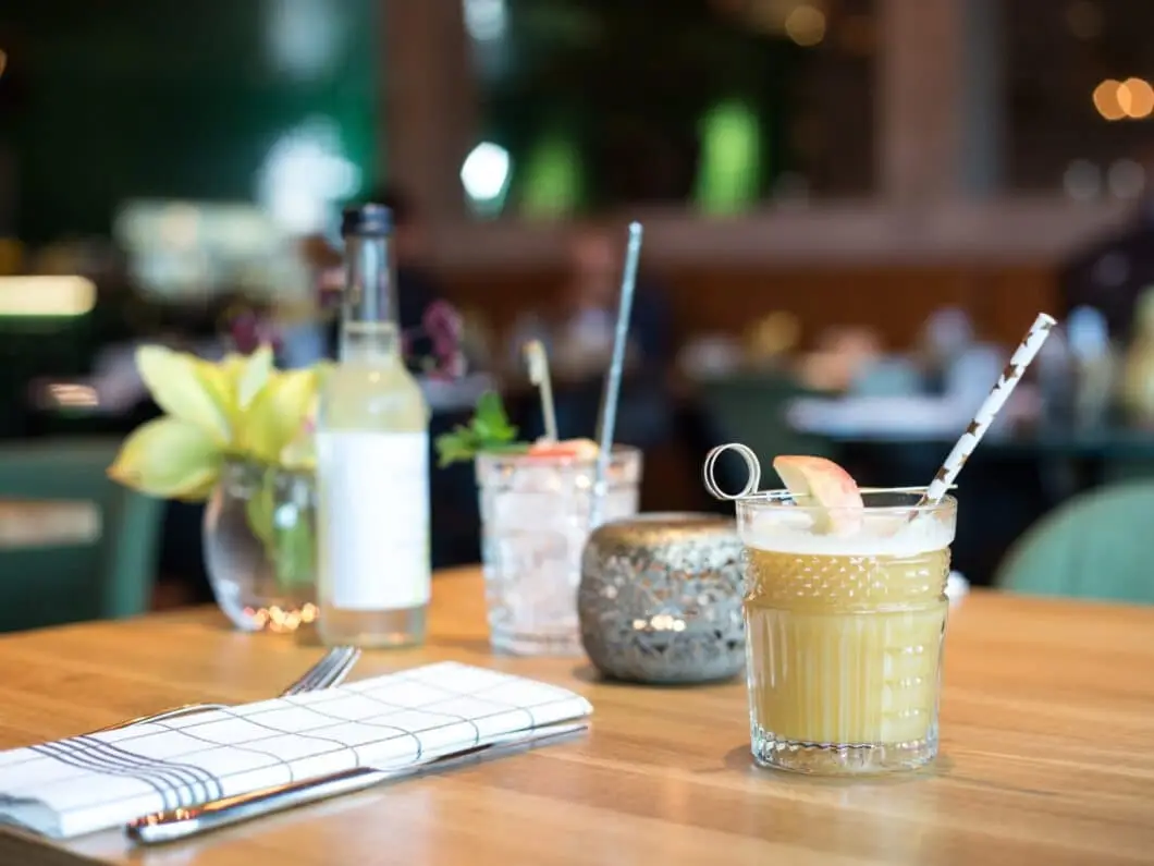A wooden table with a glass of yellow drink garnished with an apple slice, a water glass, a bottle, a napkin, and a small vase with flowers.