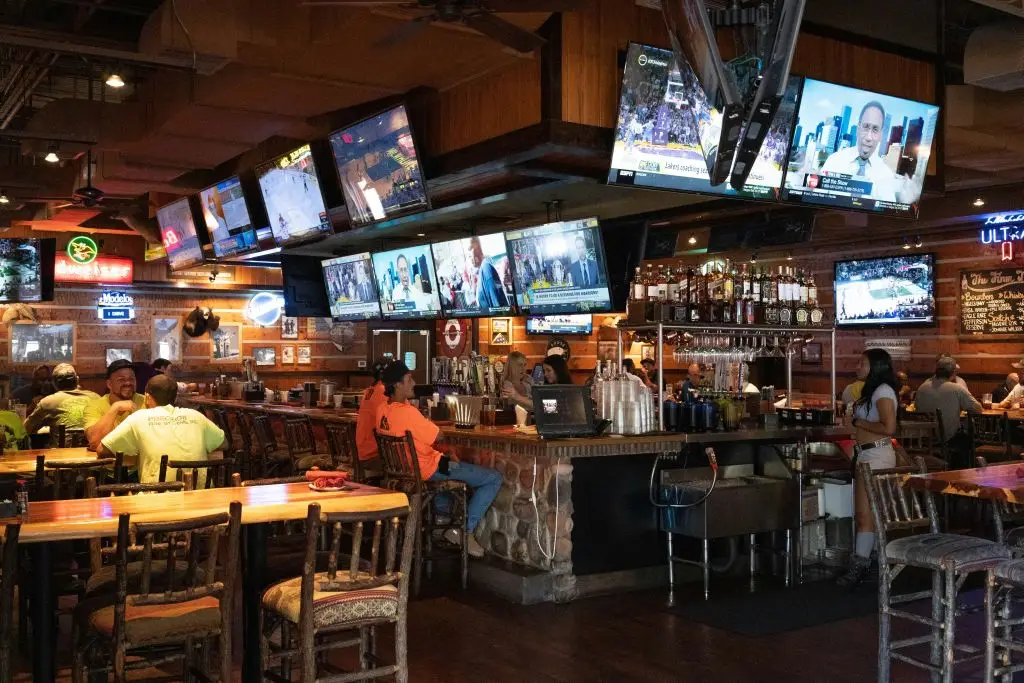 A sports bar interior with multiple televisions, a stone bar, bottles on shelves, and patrons seated at tables and the bar counter.